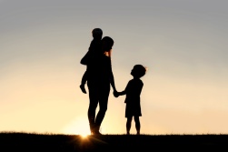 Silhouette of a young mother lovingly holding hands with her happy little child, while holding his baby brother, outside in front of a sunset in the sky.